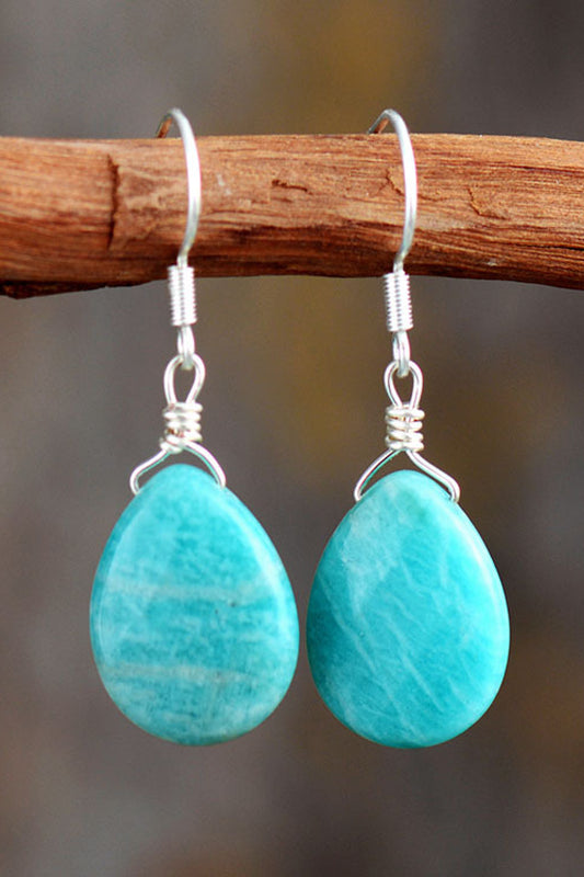 Turquoise teardrop earrings with silver hooks on a wooden branch against a blurred natural background.