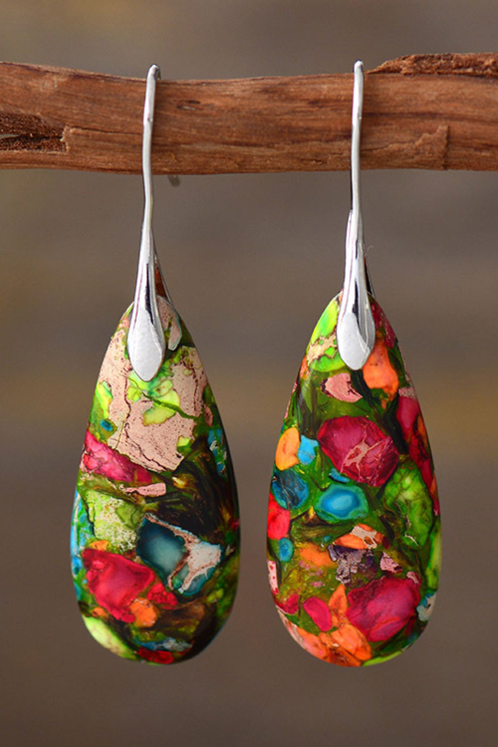 Colorful teardrop-shaped earrings hanging on a wooden stick against a brown background