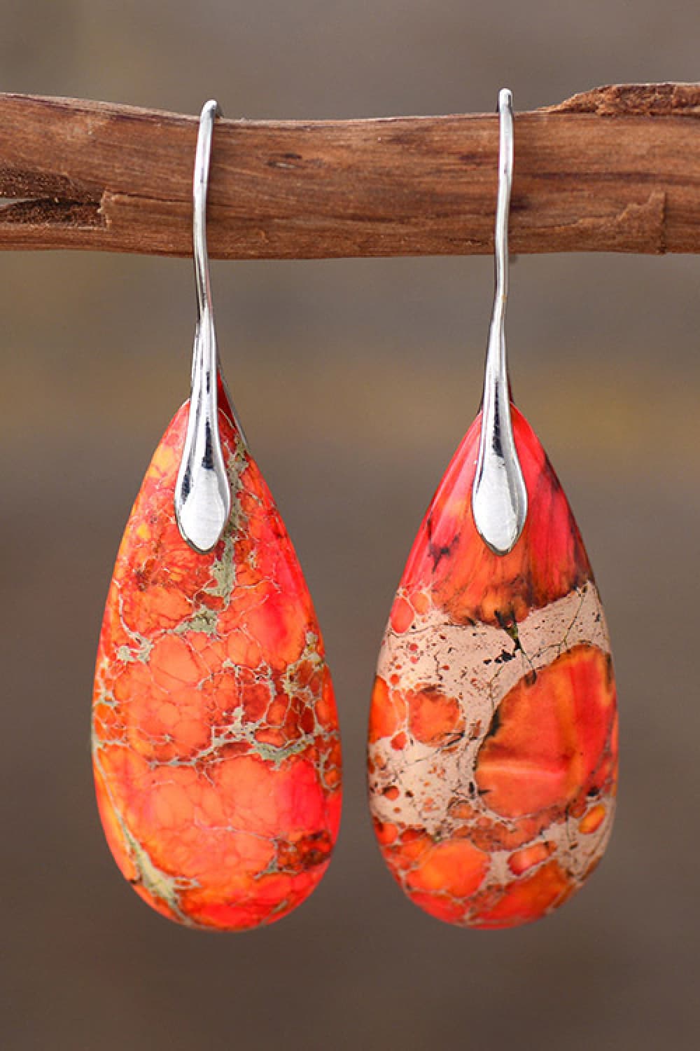 Teardrop-shaped orange earrings with silver hooks on a wooden stick against a brown background