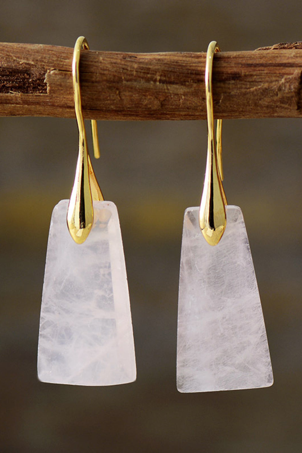 Gold earrings with white stone pendants hanging on a wooden stick against a blurred background
